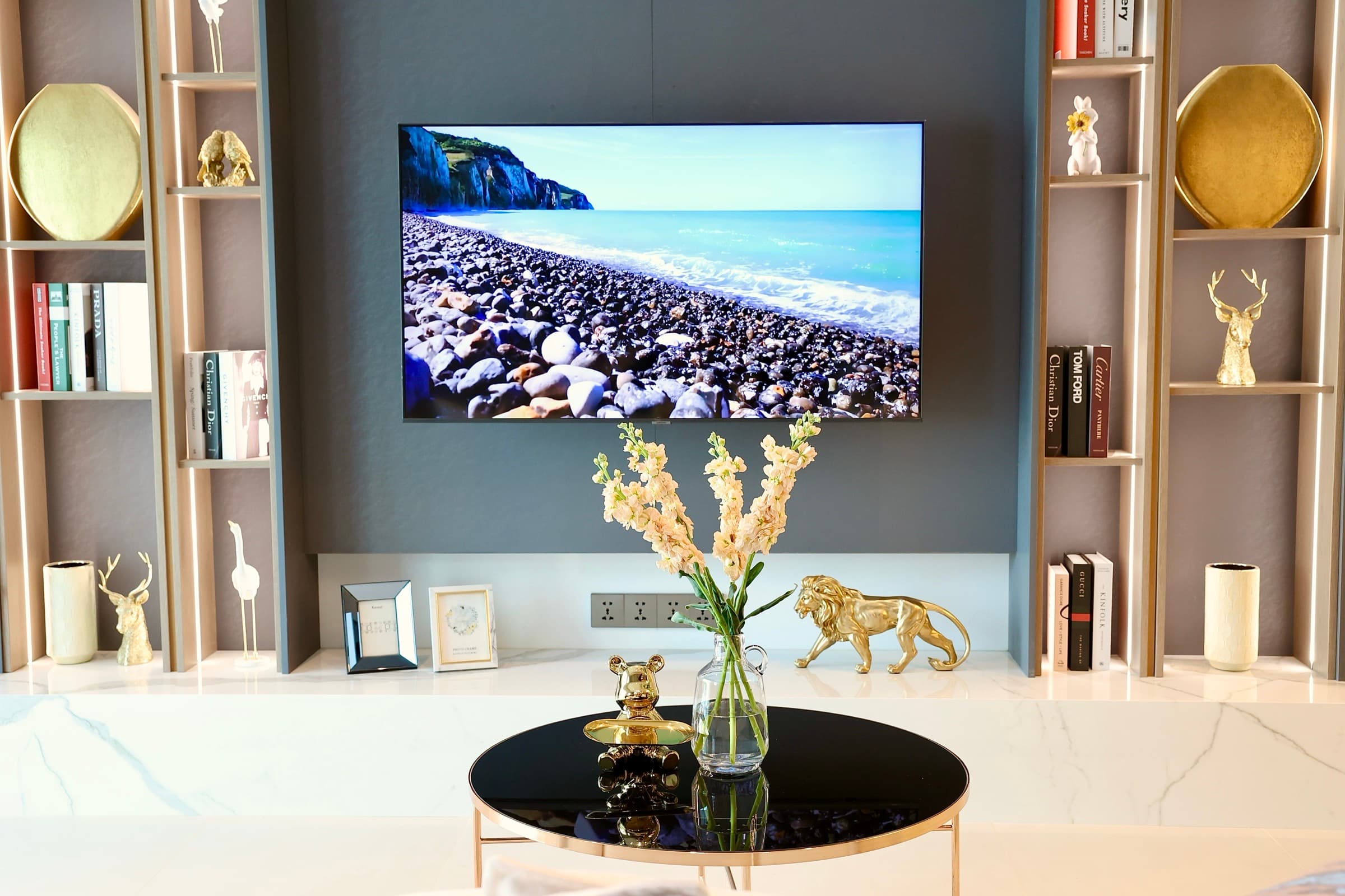 A living-room detail in Indigo — wall-mounted TV, gold deer and lion sculptures, white lilies on a marble coffee table between bookshelves.