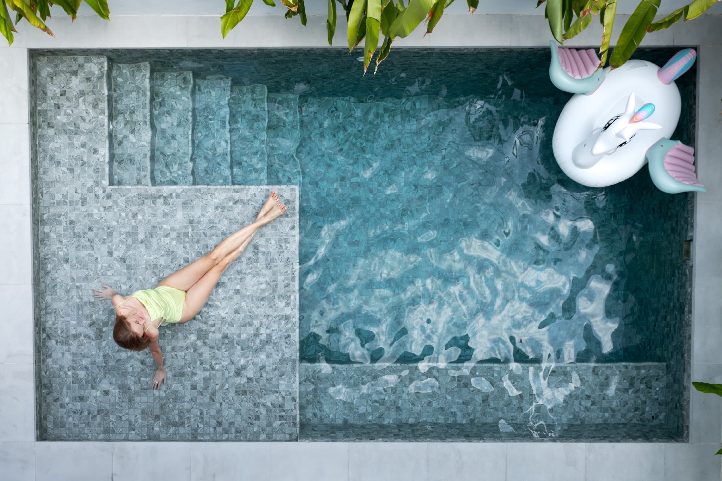 Top-down of a guest floating in the saltwater pool with a unicorn pool float on the side — slow afternoon at Villa Azure.